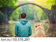 Pensive young man sitting on the pond bank with a his dog - Amazing place in Germany. Rakotzbrucke also known as Devils Bridge in Kromlau. . Стоковое фото, агентство Ingram Publishing / Фотобанк Лори