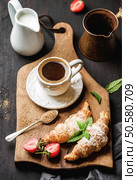 Breakfast set. Freshly baked croissants with strawberry, mint leaves and cup of coffee on wooden board served with pitcher and copper coffee pot over dark wooden backdrop, top view, selective focus. Breakfast set. Freshly baked croissants with strawberry, mint leaves and cup of coffee on wooden board over dark backdrop. Стоковое фото, агентство Ingram Publishing / Фотобанк Лори