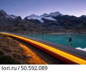 Moving modern red train near azure mountain Bianco lake, snowy rocks and purple sky at night in autumn. Bernina Express, Switzerland. Blurred train in Alps, reflection in water in fall at dusk. Стоковое фото, агентство Ingram Publishing / Фотобанк Лори