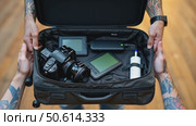 Man with tatted arm packs suitcase for adventure travel trip. Packing bag with camera, laptop, portable battery charger. Modern travel business concept. Стоковое AI-видео, аниматор Sergey Grishin / Фотобанк Лори