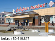 Супермаркет «Северное сияние», город Магадан, Магаданская область, Россия. На улице перед торговым центром молодые люди и девушки кормят голубей. На площади около здания магазина и Сбербанка установлена скульптура «Один червонец». Повседневная жизнь в Сибири и на Дальнем Востоке России. Весенний сезон. Май. Редакционное фото, фотограф Андрей Степанов / Фотобанк Лори