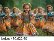A bright photo of girls in traditional Russian folk costumes, holding hands in a round dance in nature. In the center of the frame is a smiling girl with pigtails, decorated with a wreath of artificial flowers. Her outfit is richly embroidered with patterns, fringe and lace, in rich shades of yellow, blue and orange. In the background are other children in similar costumes, creating an atmosphere of a folk festival, fun and children's joy against the backdrop of a green forest or field. The photo conveys the spirit of Slavic traditions, the purity of childhood and cultural heritage. Стоковое AI-изображение, AI-художник Анна Кривицкая / Фотобанк Лори