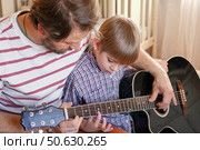Handsome dad teaches his cute son to play the guitar sitting on sofa in the living room at home. Happy Father's Day. Father teaching his child to play guitar. Kid Learning music skill. Art education. Стоковое фото, фотограф Марина Демидюк / Фотобанк Лори