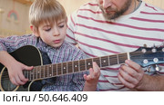 Handsome dad teaches his cute son to play the guitar sitting on sofa in the living room at home. Happy Father's Day. Father teaching his child to play guitar. Kid Learning music skill. Art education. Стоковое видео, видеограф Марина Демидюк / Фотобанк Лори