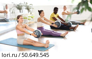 Sporty young girl enjoying Pilates workout with diverse group in sunlit studio, sitting on mat and doing overhead ball lift exercise with straight arms, promoting shoulder strength and core stability. Стоковое видео, видеограф Яков Филимонов / Фотобанк Лори