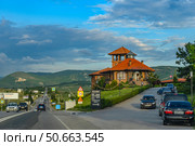 A modern cafe with a red tile roof against the backdrop of picturesque Crimean hills and green valleys. The building in the style of Mediterranean architecture rises on the side of the highway leading deep into the mountainous Crimea. In the foreground is a parking lot with cars and a sign with the establishment's menu. Soft evening light emphasizes the atmosphere of coziness and hospitality, creating an ideal scene for travelers. Редакционное фото, фотограф Анна Кривицкая / Фотобанк Лори