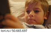 Cute kid boy in pajamas lying in bed watching smartphone, smiling. Children's screen time. Child plays games using phone at the evening or night. Social media addiction. Childhood, modern technology. Стоковое видео, видеограф Марина Демидюк / Фотобанк Лори