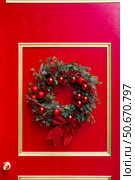 Christmas Eve. A Christmas wreath on a red front door. Christmas decorations in the interior. The concept of preparing a house for Christmas and New Year. Стоковое фото, фотограф Photojuli / Фотобанк Лори