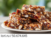 Close-up of nut and dried fruit brittle bars stacked on a white plate, glistening with caramelized syrup, against a blurred green background. Стоковое AI-изображение, AI-художник Катерина Бондарец / Фотобанк Лори