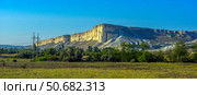 White Rock (Ak-Kaya) in Crimea is a monumental limestone formation with vertical cliffs, towering over a green valley. The rocky massif is flooded with sunlight, emphasizing the texture and light cream shade of the limestone. In the foreground are meadows and trees, creating a picturesque natural contrast with the majestic relief of the mountains. The sky is clear, bright blue, which adds a sense of space and grandeur to the frame. Стоковое фото, фотограф Анна Кривицкая / Фотобанк Лори