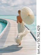 Elegant woman in flowing white dress walking on tropical boardwalk holding a parasol, surrounded by turquoise water under soft sunlight, viewed from behind. Стоковое AI-изображение, AI-художник Катерина Бондарец / Фотобанк Лори