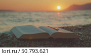 Open Bible book on pebbles with cross pendant during tranquil beach sunset. Стоковое видео, видеограф Igor Tichonow / Фотобанк Лори