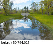 Beautiful deserted park in spring. View of stone pedestrian bridge with mirror reflection in quiet river. Green beds with dandelions and fresh young foliage on trees. Picturesque landscape of sunny May day. Стоковое фото, фотограф Виктория Катьянова / Фотобанк Лори