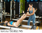 Girl trainer helps young woman to perform exercises in lying position on pilates reformer. Стоковое фото, фотограф Яков Филимонов / Фотобанк Лори