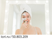 Cheerful woman with a towel on her hair and in a clay face mask fooling around with cucumbers in her hands. Taking care of beauty at home. Стоковое фото, фотограф Михаил Решетников / Фотобанк Лори