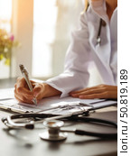 Female doctor writing on clipboard with stethoscope on desk. Excellent for healthcare articles, medical branding, or professional clinic content. Стоковое AI-изображение, AI-художник Александра Троицкая / Фотобанк Лори