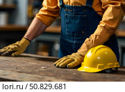 A worker in a blue jumpsuit stands at a desk with a yellow helmet and gloves. Стоковое AI-изображение, AI-художник Галина Эмберг / Фотобанк Лори