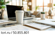 Mock up paper coffee cup with cup holder on a wooden table nearby scattered documents and an open laptop as morning light fills the office. Copy space. Стоковое AI-изображение, AI-художник Юлия Ильина / Фотобанк Лори