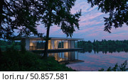 Minimalist floating home hovering above tranquil lake waters, glass walls mirroring golden sunset light amid dense green shoreline vegetation. Стоковое AI-изображение, AI-художник Данил Руденко / Фотобанк Лори