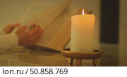 Burning candle in foreground with person reading a large book behind indoors. Стоковое видео, видеограф Igor Tichonow / Фотобанк Лори