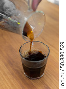 A person pours dark coffee from a glass carafe into a clear glass cup on a wooden table. The coffee has a rich, dark color and a frothy top. Стоковое фото, фотограф PAv / Фотобанк Лори