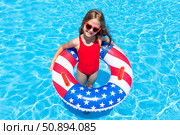 Top view happy little girl in red swimsuit and sunglasses stands in outdoor swimming pool with a lifebuoy decorated with American flag print. Vacation in USA concept. 4th of July pool party. Стоковое фото, фотограф Ирина Михайличенко / Фотобанк Лори
