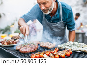Senior Caucasian man with a beard grilling meat and vegetables outdoors. Стоковое AI-изображение, AI-художник Евгения Пархоменко / Фотобанк Лори