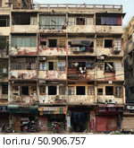 A photograph of an old apartment building in an Asian metropolis, with a facade covered in traces of time and urban bustle. Worn walls, various windows, clothes on the lines, air conditioners, satellite dishes and a chaotic arrangement of elements give the building a characteristic atmosphere of a densely populated area. Below are small shops with signs in the local language and parked motor scooters. This visual image perfectly conveys everyday life, urban density and architecture of developing countries. Стоковое AI-изображение, AI-художник Анна Кривицкая / Фотобанк Лори