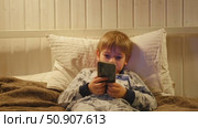 Cute kid boy in pajamas lying in bed watching smartphone, smiling. Children's screen time. Child plays games using phone at the evening or night. Social media addiction. Childhood, modern technology. Стоковое видео, видеограф Марина Демидюк / Фотобанк Лори