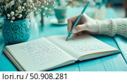 Contemplative journaling: Hand holding a pen writing in a notebook on a turquoise table with a flower vase, capturing a moment of inspiration. Стоковое AI-изображение, AI-художник Данила Большаков / Фотобанк Лори