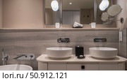 Bathroom. Beige and cream shades. Two round white wash basins. There's a soap dish, a box of napkins on the countertop. Metal faucets on a tiled wall. The lamps are lit. Reflection in the wall mirror. Стоковое фото, фотограф Вера Ивановна Кубарева / Фотобанк Лори