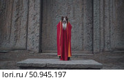 Old priest with a long white beard and dressed in red and gold robes is praying with his hands joined in front of the monolithic rock hewn Church of Lalibela, a UNESCO World Heritage site. Стоковое AI-видео, аниматор Данил Руденко / Фотобанк Лори