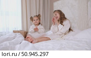 Two little girls dressed in elegant white dresses sit on a bed in the morning, joyfully clapping their hands and celebrating a festive occasion. Childhood, happiness concept. High quality footage. Стоковое видео, видеограф Ирина Михайличенко / Фотобанк Лори