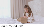 Child, little girl in white dress is sitting on a bed with white bedding, opening gift box wrapped in kraft paper. Holidays concept. High quality footage. Стоковое видео, видеограф Ирина Михайличенко / Фотобанк Лори