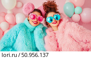 Vibrant celebration with two cheerful young women in playful faux fur sweaters and novelty heart-shaped eye-wear, surrounded by pastel balloons, capturing joyous friendship moments, suitable for a ha. Стоковое AI-изображение, AI-художник Данила Большаков / Фотобанк Лори