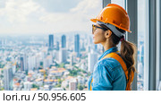 Professional female engineer in orange hard hat observes sprawling cityscape from high-rise, symbolizing urban development and ambition. Стоковое AI-изображение, AI-художник Данила Большаков / Фотобанк Лори