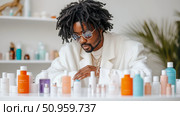 A discerning man with dreadlocks and round sunglasses thoughtfully examines an array of vibrant skincare products on a pristine white counter, conveying modern grooming and self-care. Стоковое AI-изображение, AI-художник Данила Большаков / Фотобанк Лори