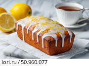 Homemade lemon drizzle cake with icing. Classic lemon pound cake with citrus glaze and zest. Whole loaf, side view, close up. Fresh homemade baked dessert with lemon slices. Blurred tea set background. Стоковое AI-изображение, AI-художник Марина Демидюк / Фотобанк Лори
