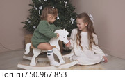 Two sisters have fun and play at home near the Christmas tree. The older sister sits on a floor, rocking a wooden rocking horse, while the younger toddler sister sits happily on it. Стоковое видео, видеограф Ирина Михайличенко / Фотобанк Лори