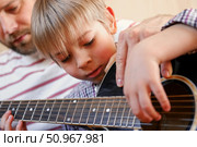 Handsome dad teaches his cute son to play the guitar sitting on sofa in the living room at home. Happy Father's Day. Father teaching his child to play guitar. Kid Learning music skill. Art education. Стоковое фото, фотограф Марина Демидюк / Фотобанк Лори