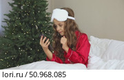 A little girl in red pajamas with a white sleep mask on her head sits on a bed on Christmas morning near decorated tree and uses smartphone, writes message to friends, parents or plays online game. Стоковое видео, видеограф Ирина Михайличенко / Фотобанк Лори