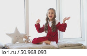 Happy laughing little girl in a red festive dress sitting near big window on Christmas morning, looking at camera, saying hello and waves her hands in greeting. High quality footage. Стоковое видео, видеограф Ирина Михайличенко / Фотобанк Лори