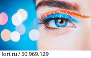 Mesmerizing close-up of a blue eye with vibrant, artistic blue and orange makeup, enhanced by sparkling white accents, set against a dreamy blue bokeh background. Стоковое AI-изображение, AI-художник Данила Большаков / Фотобанк Лори