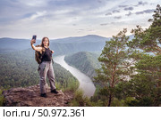A hiker stands atop a rocky ledge, capturing a selfie against a breathtaking backdrop of lush green forests and a winding river. A female solo traveler takes a selfie photo on a mountain. Стоковое фото, фотограф Владимир Разгуляев / Фотобанк Лори