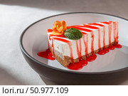 Cheesecake slice with graham crust, strawberry syrup, mint leaf and edible flower served on a modern ceramic plate in soft natural light. Стоковое фото, фотограф Aleksandr Gavrilychev / Фотобанк Лори