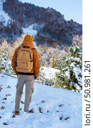 Rear view of a man in a red jacket and hat with a yellow backpack admiring a beautiful mountain view with an autumn-winter forest landscape Hiking and tourism. Стоковое фото, фотограф Елена Гоосен / Фотобанк Лори