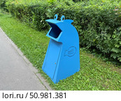 A bright blue, cartoon-like trash can with fish decorations sits on a sidewalk beside a grassy area and bushes. Concept of: Public space cleanliness. Стоковое фото, фотограф Ольга Петрова-Апостолова / Фотобанк Лори