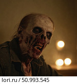 A decayed zombie man sits in a shadowy room, his skeletal face and lifeless eyes capturing the essence of horror. Flickering candles illuminate his grotesque features Generative AI. Стоковое AI-изображение, AI-художник Юлия Петровская / Фотобанк Лори