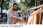 Man browsing secondhand clothes at an outdoor market on a sunny day, enjoying a casual shopping experience surrounded by racks of colorful garments and greenery. Стоковое AI-изображение, AI-художник Катерина Бондарец / Фотобанк Лори