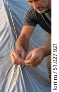 Camper adjusting tensioning rope on tent during camping trip. Стоковое AI-изображение, AI-художник Юрий Поздников / Фотобанк Лори